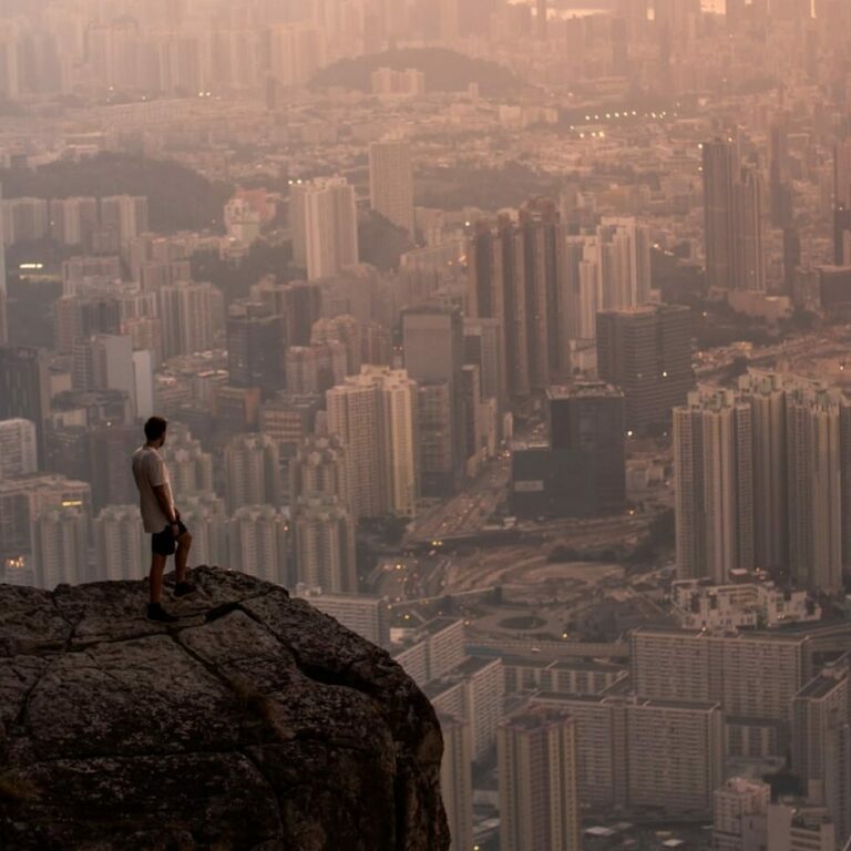 man sitting on rock looking at city skyline during daytime
